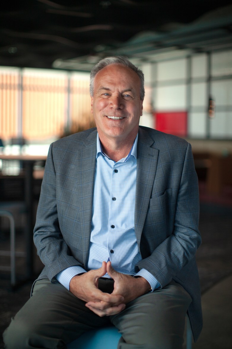 A person in a blazer and collared shirt sits indoors with hands clasped and smiles toward the camera.