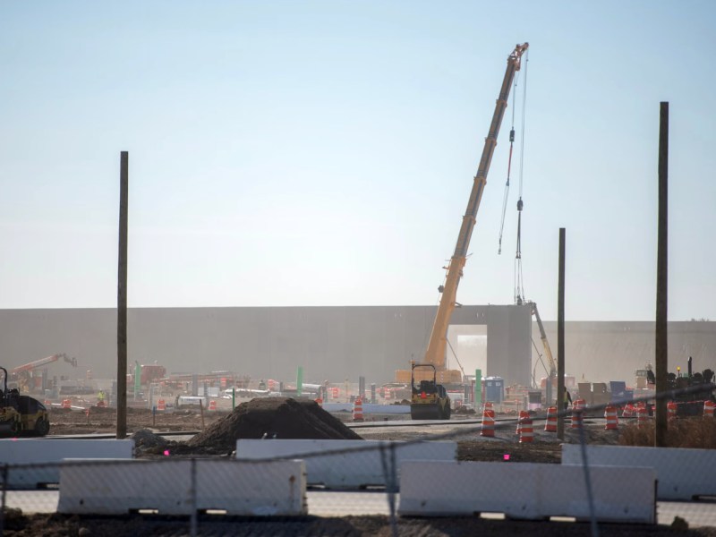 A large yellow crane's lifting line is attached to a large concrete wall panel at a construction site with rollers, dirt piles, traffic barrels, and vertical posts behind a fence.