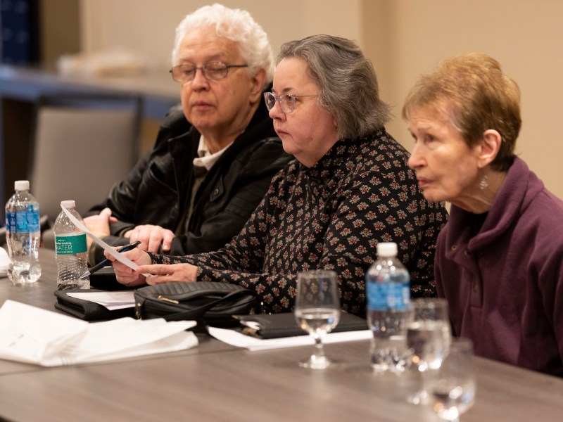 Three people sit at a table with papers, notebooks and water bottles in front of them.