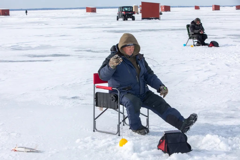 A person in a hooded winter jacket sits on a folding chair on a frozen body of water and holds a fishing line, with ice shanties and a vehicle in the distance.