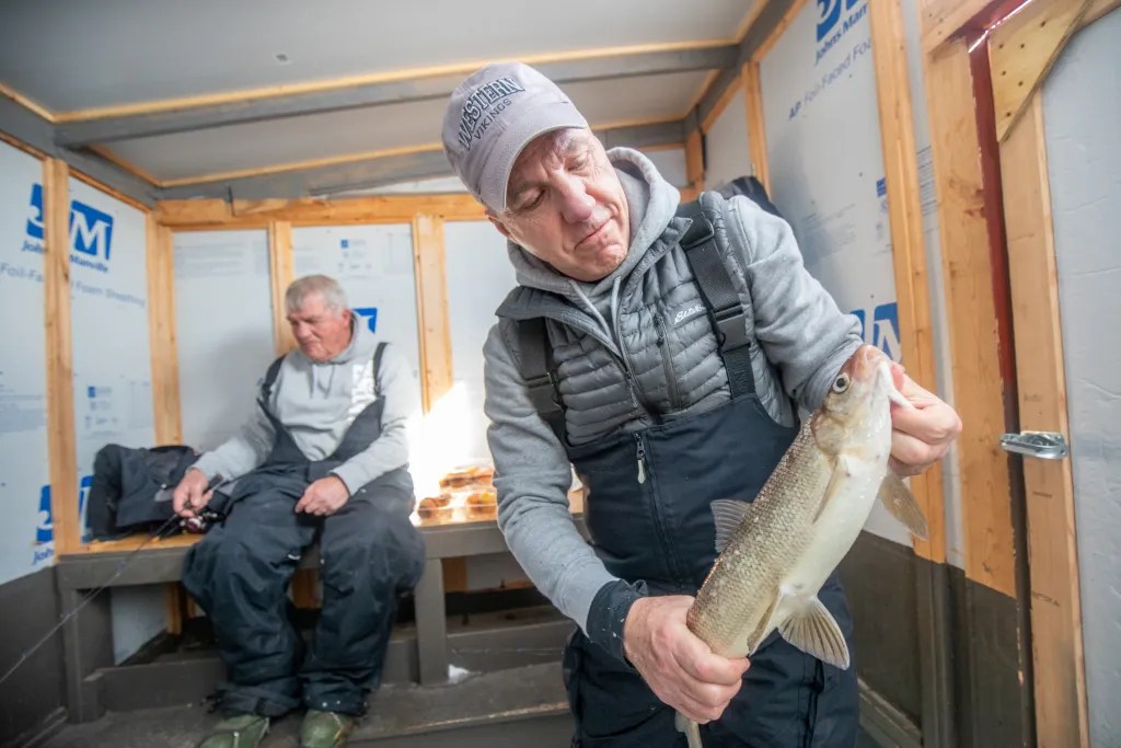 A person in insulated overalls holds a fish inside an ice fishing shelter, while another person sits on a bench in the background holding a fishing rod.