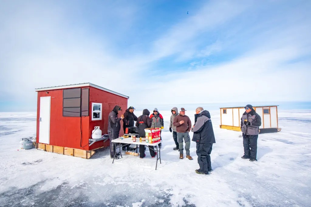 People in winter jackets stand on a frozen body of water beside a red ice fishing shanty and a folding table with food and supplies, with another shanty nearby.