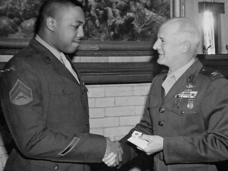 Two people in military uniforms shake hands, with one holding a small card and a painting of soldiers in combat hanging on the wall behind them.