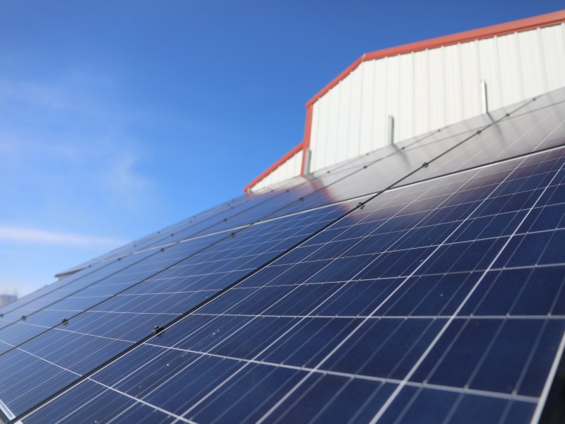 Solar panels reflect sunlight beside a white metal building with a red roof under a blue sky.