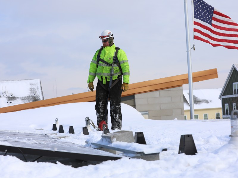 Man in yellow jacket stands on snow-covered roof next to solar panel and American flag.