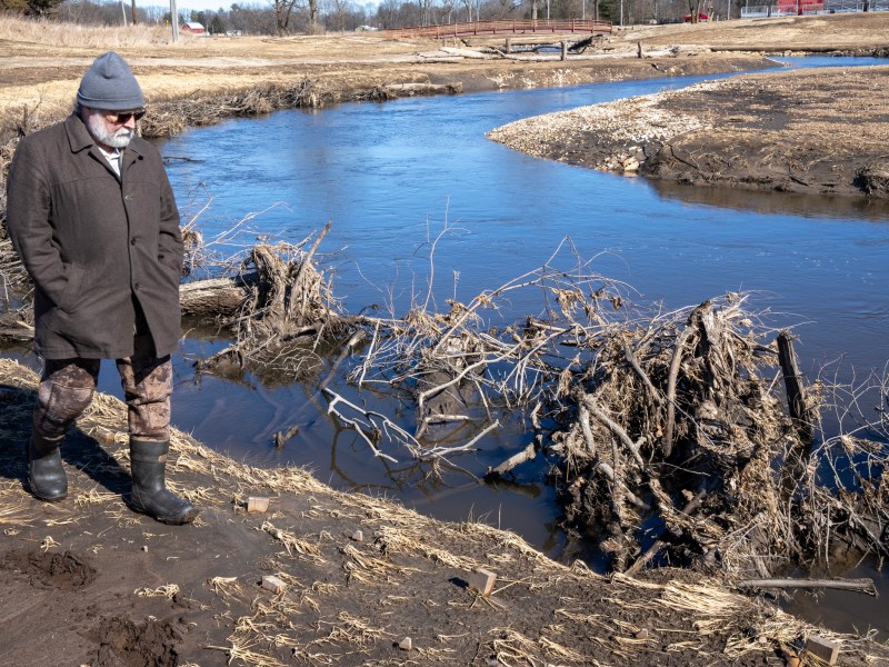 Man in coat, hat, sunglasses and rubber boots walks past a creek.