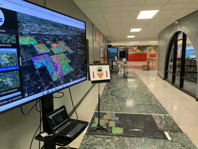 Large display screen shows a digital map with colored areas beside a hallway with a sign reading "CAFO proposed" and red lockers in the background.